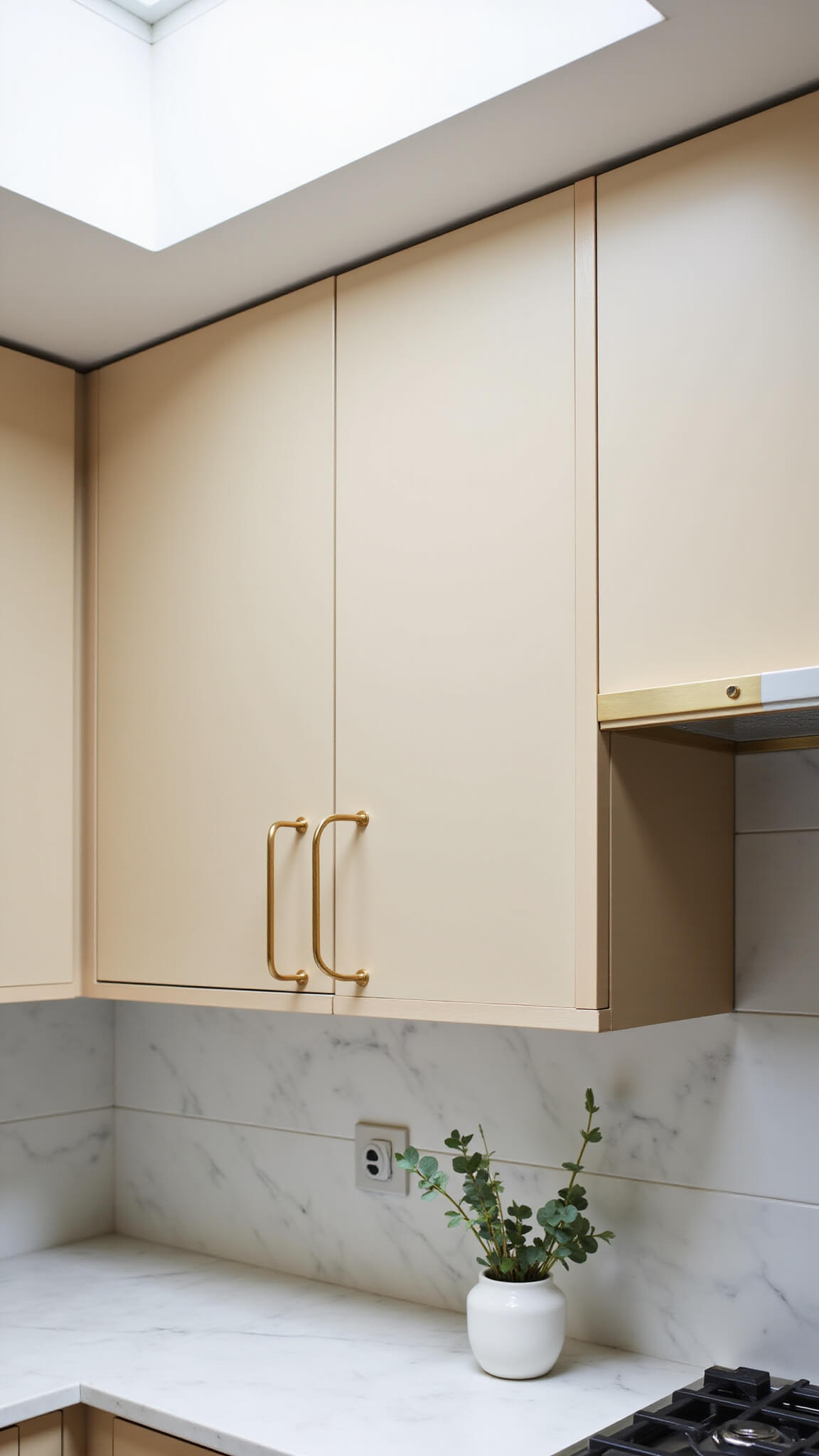 Close-up of modern brass cabinet pulls on beige flat-panel kitchen cabinets under skylight, with marble backsplash and white vase with eucalyptus.
