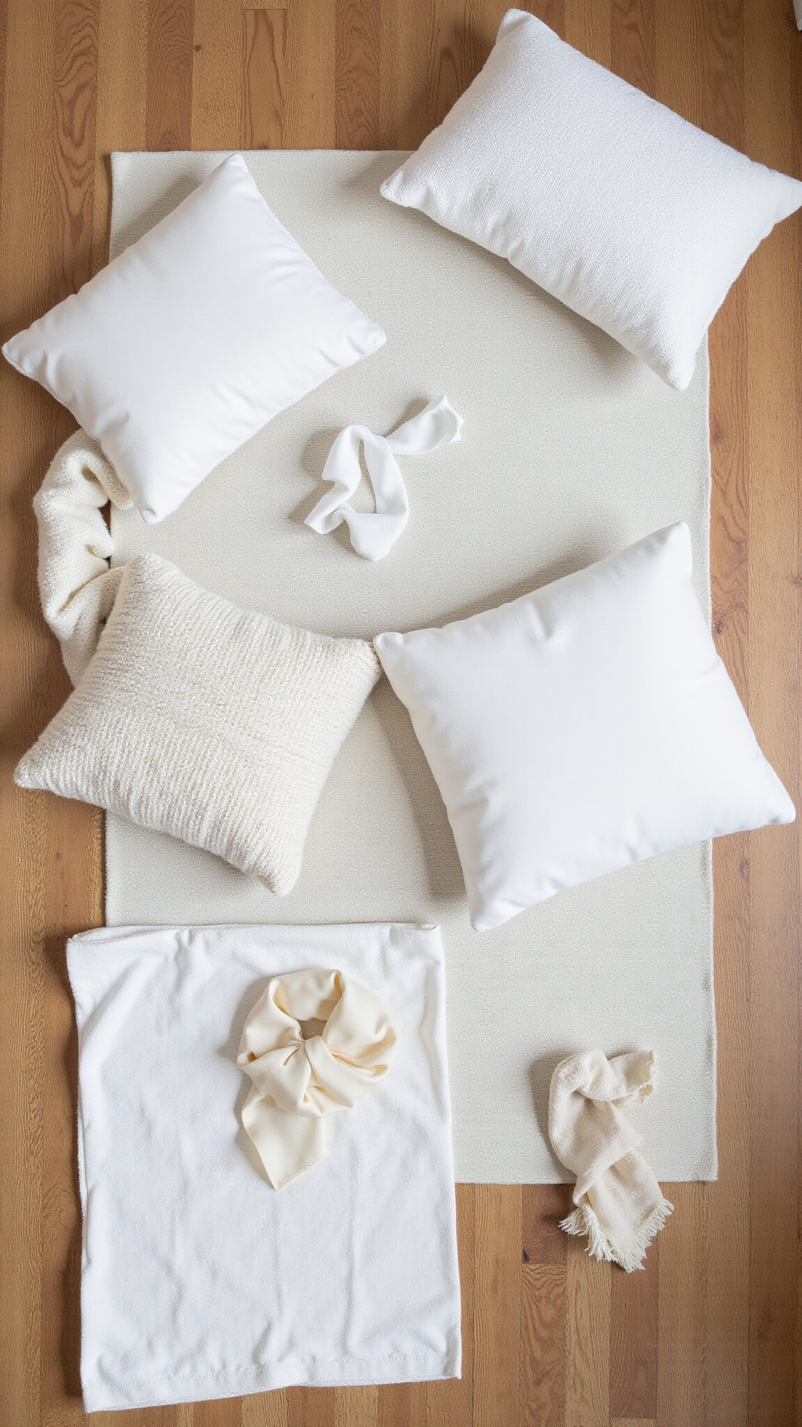 Flat-lay of layered white bedroom textiles on hardwood floor with pillows, blankets, and rug in varied white tones.
