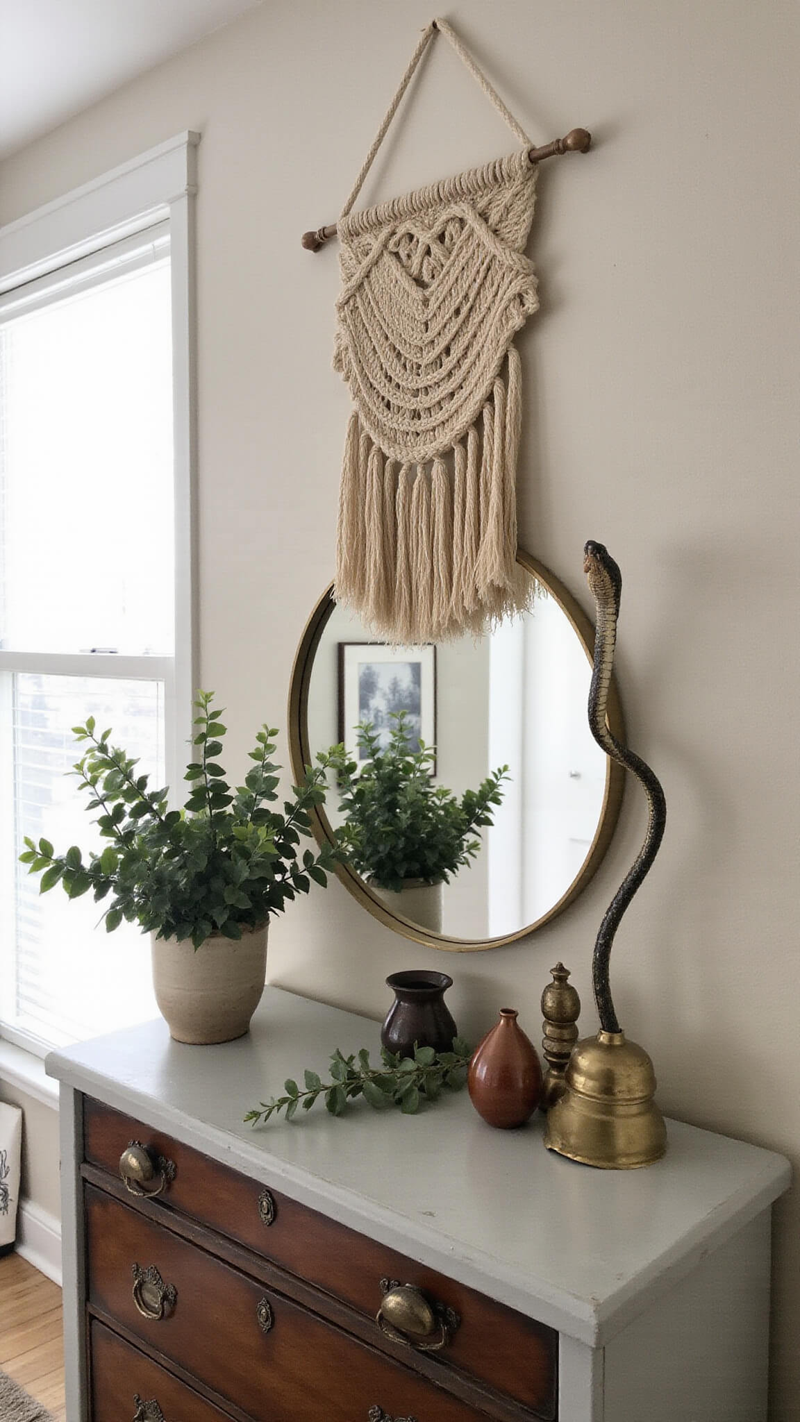 Handwoven wall hanging above antique dresser with brass mirror, ceramic vessels, eucalyptus, and snake plant in moody morning light.