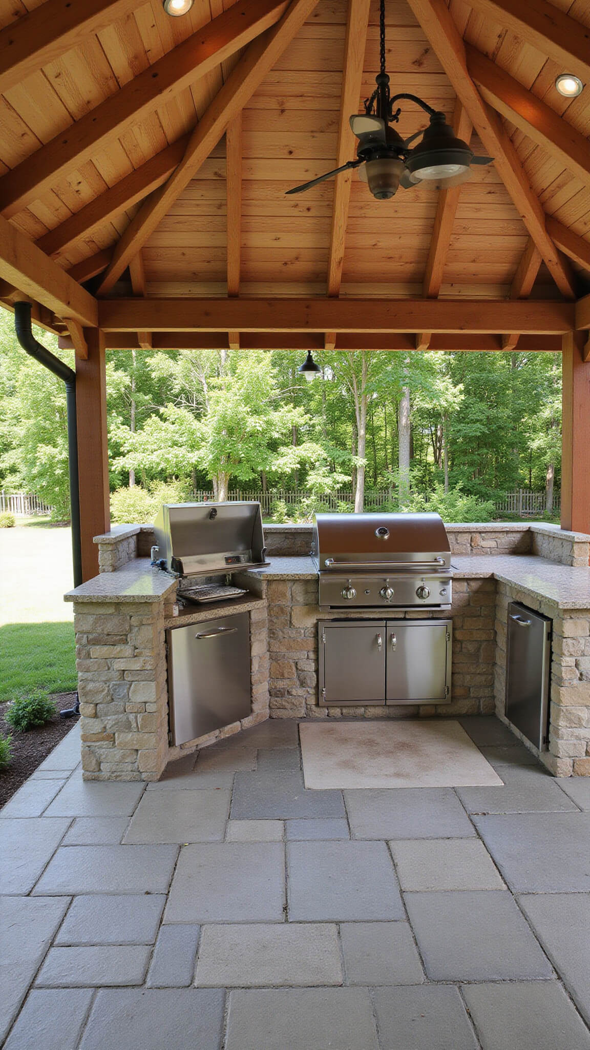 Outdoor kitchen gazebo with cedar shake roof, stone base, stainless steel grill, granite counters, pendant lights, bar seating, and surrounding herb garden under pergola.