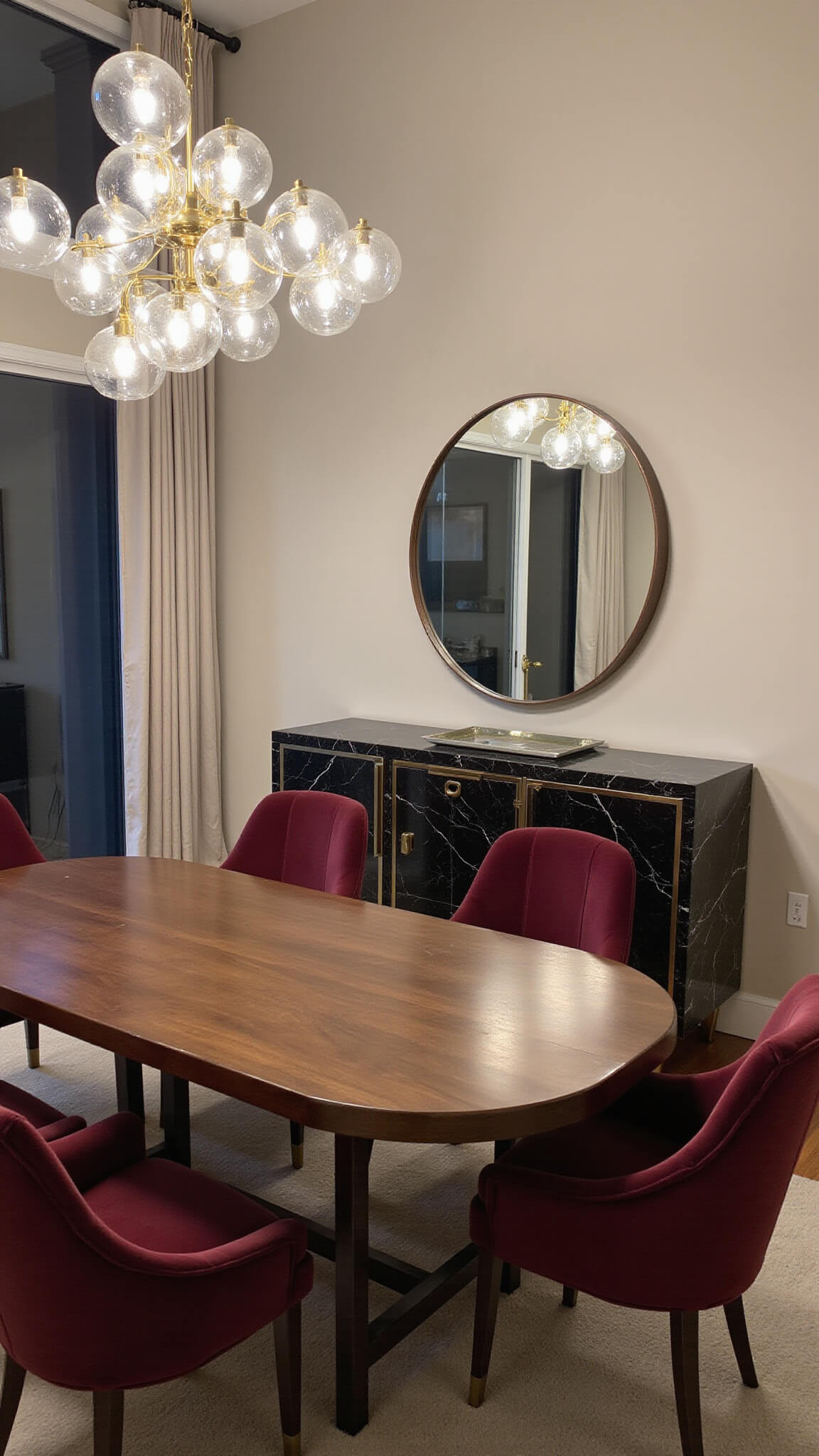Contemporary dining room with sculptural brass chandelier over oval walnut table, burgundy velvet chairs, floor-to-ceiling windows, and black marble buffet.