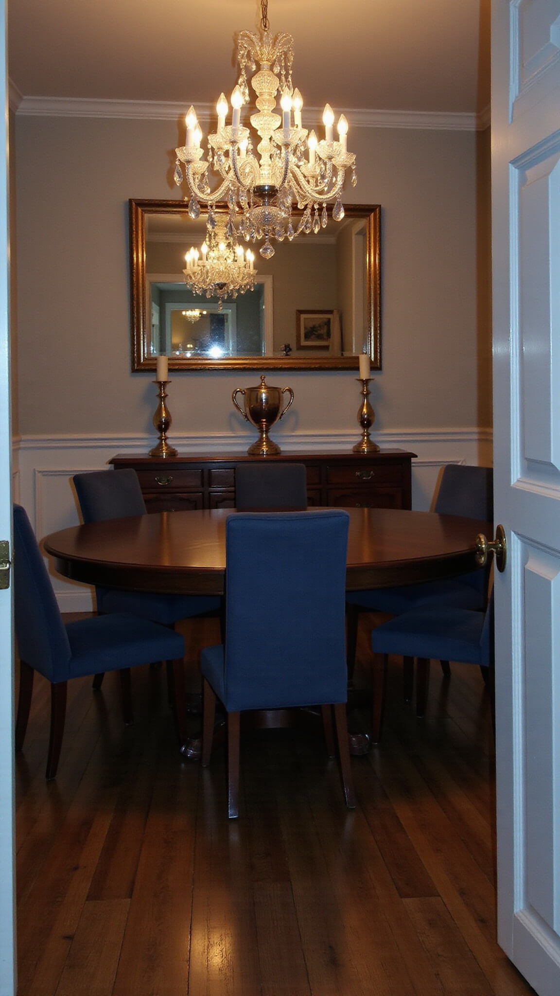 Elegant formal dining room at dusk with dark oak round table, slate blue velvet chairs, crystal chandelier, silver candelabras, and gilt-framed mirror, viewed wide-angle from doorway.