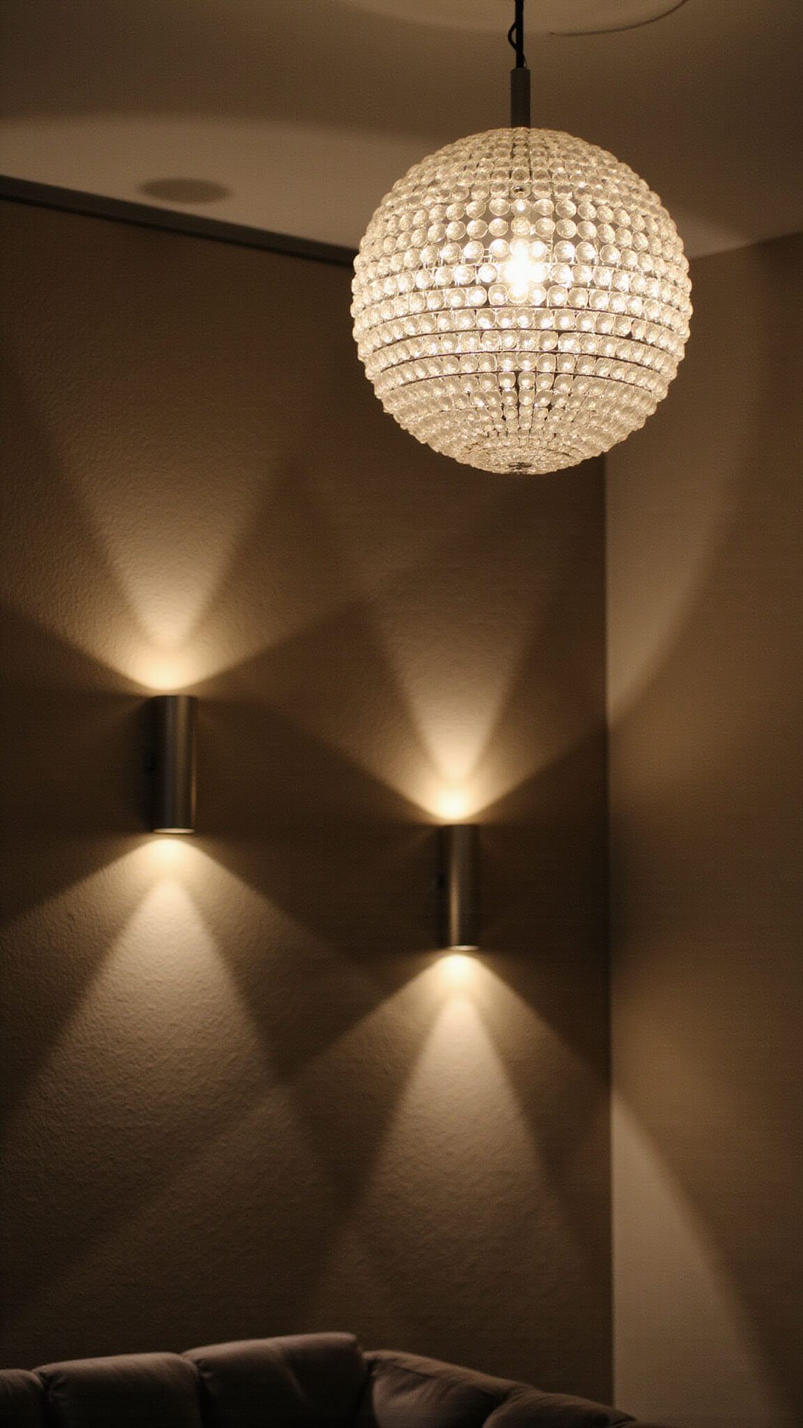 Close-up of layered nursery lighting at night with chandelier, wall sconces, and LED strips highlighting textured grasscloth wallpaper, velvet upholstery, and champagne metallic accents.