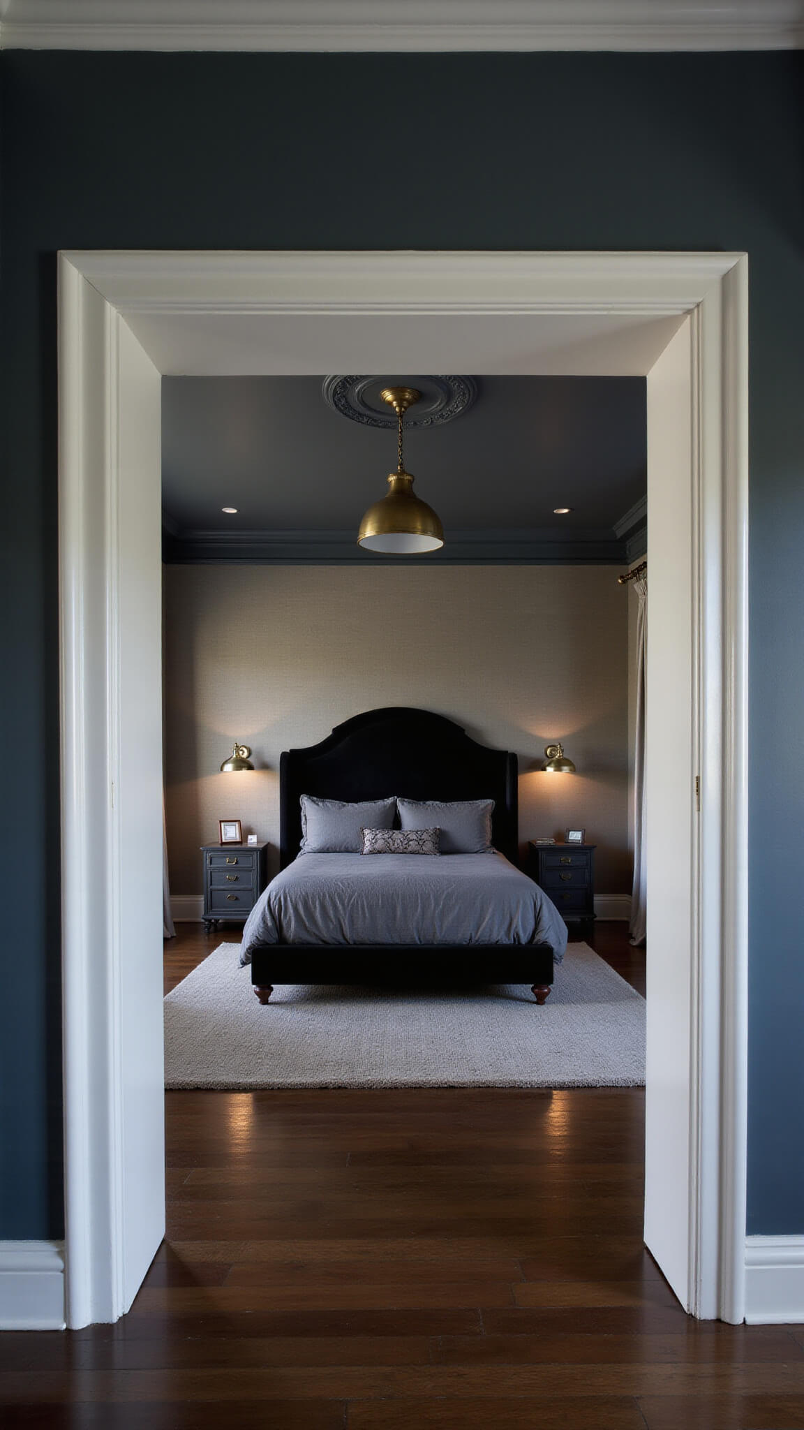 Moody master bedroom at dusk with charcoal linen king bed, black velvet headboard, brass pendant lights, grasscloth accent wall, silvery silk drapes, and dark hardwood floors.