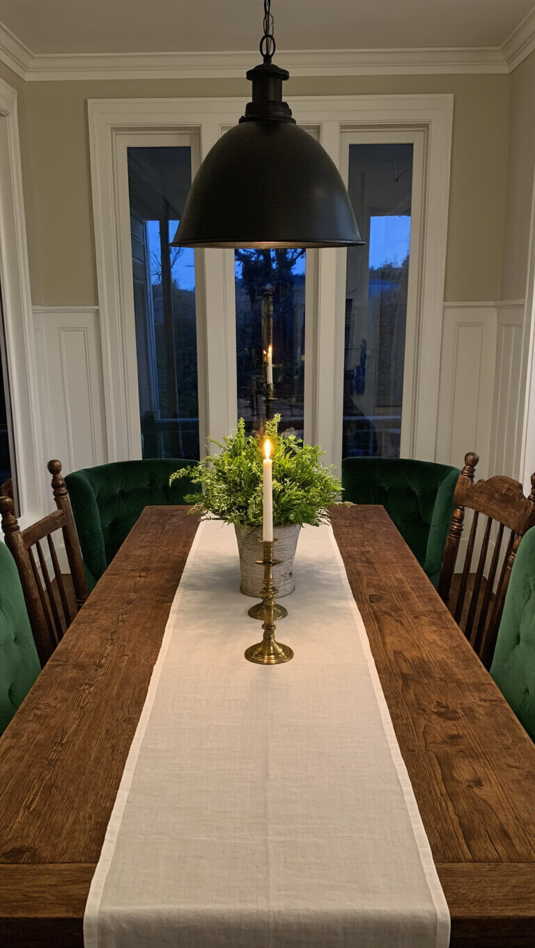 Twilight dining room with vintage pine table set for six, velvet and wood chairs, aged brass candlesticks, and black iron pendant light in cozy, symmetrical setting.