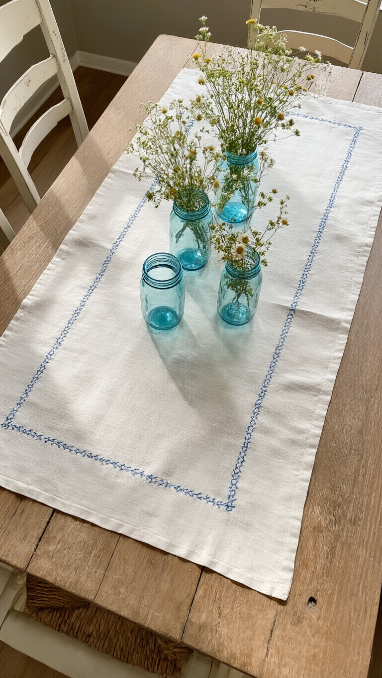 Overhead view of rustic harvest table with vintage cream and blue embroidered linens, wildflowers in antique blue jars, and distressed white chairs.