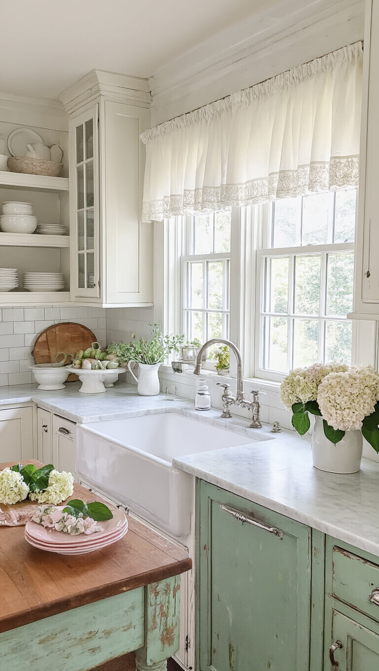Vintage-style 12x14 kitchen with worn white cabinets, sage green island, marble counters, farmhouse sink under window with cafe curtains, and open shelves displaying white ironstone and pink depression glass.
