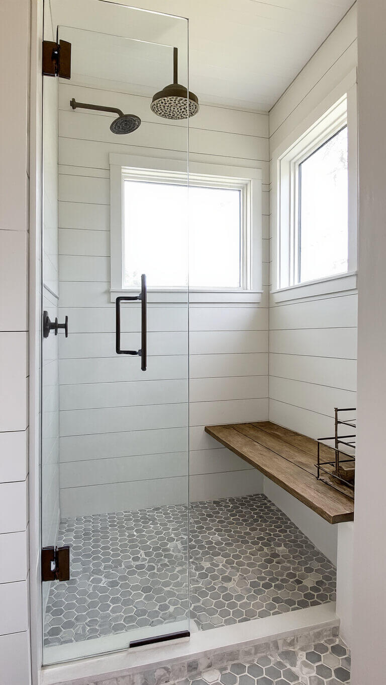 Modern farmhouse shower with shiplap-style porcelain walls, hex tile floor, frameless glass, oil-rubbed bronze fixtures, reclaimed wood bench, and aged brass corner caddy.
