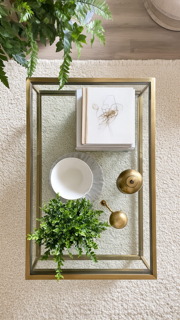 Overhead view of styled brass and glass coffee table with art books, white bowl, brass decor, and greenery on neutral rug in soft morning light.