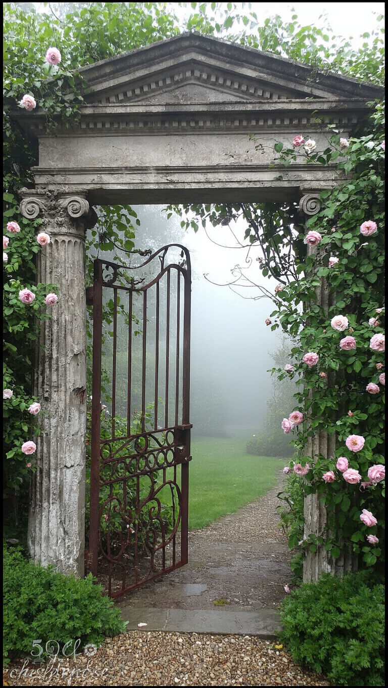 Weathered stone and rusted iron garden entrance made from salvaged columns and pediment, framed by climbing roses and clematis in morning fog.