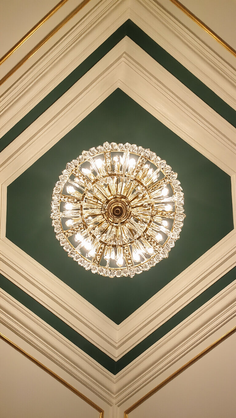 Upward shot of ornate ceiling with plaster medallion, tiered brass and crystal chandelier, geometric cream molding on hunter green background, and gold leaf accents highlighted by dramatic lighting.
